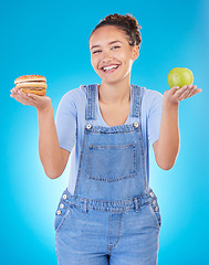 Image showing Happy, portrait and woman with burger, apple and choice in studio isolated on a blue background. Smile, fruit and person with sandwich, fast food or comparison for healthy diet, nutrition or wellness