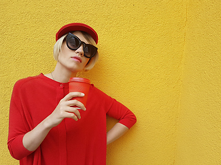 Image showing Female model in long red sweater and red hat with red paper cup standing on yellow background. Outdoor photo of beautiful caucasian woman in sweater drinks coffee in front of cafe