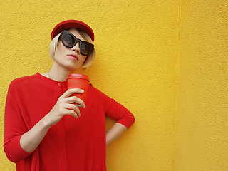 Image showing Female model in long red sweater and red hat with red paper cup standing on yellow background. Outdoor photo of beautiful caucasian woman in sweater drinks coffee in front of cafe