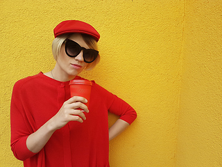 Image showing Female model in long red sweater and red hat with red paper cup standing on yellow background. Outdoor photo of beautiful caucasian woman in sweater drinks coffee in front of cafe