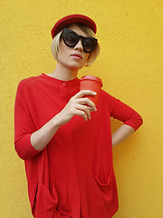 Image showing Female model in long red sweater and red hat with red paper cup standing on yellow background. Outdoor photo of beautiful caucasian woman in sweater drinks coffee in front of cafe