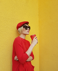 Image showing Female model in long red sweater and red hat with red paper cup standing on yellow background. Outdoor photo of beautiful caucasian woman in sweater drinks coffee in front of cafe