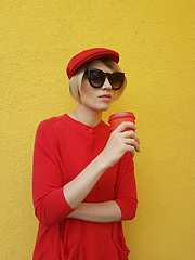 Image showing Female model in long red sweater and red hat with red paper cup standing on yellow background. Outdoor photo of beautiful caucasian woman in sweater drinks coffee in front of cafe