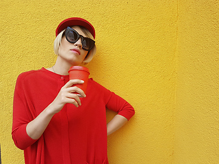 Image showing Female model in long red sweater and red hat with red paper cup standing on yellow background. Outdoor photo of beautiful caucasian woman in sweater drinks coffee in front of cafe