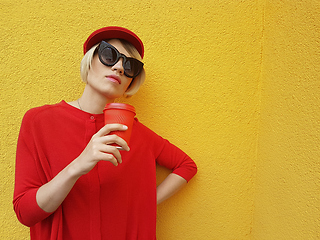 Image showing Female model in long red sweater and red hat with red paper cup standing on yellow background. Outdoor photo of beautiful caucasian woman in sweater drinks coffee in front of cafe