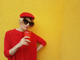 Image showing Female model in long red sweater and red hat with red paper cup standing on yellow background. Outdoor photo of beautiful caucasian woman in sweater drinks coffee in front of cafe