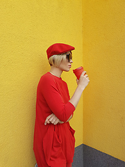 Image showing Female model in long red sweater and red hat with red paper cup standing on yellow background. Outdoor photo of beautiful caucasian woman in sweater drinks coffee in front of cafe