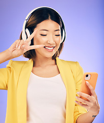 Image showing Peace sign, selfie and happy Asian woman in studio smile for social media, online post or mobile app. Headphones, music and female person with emoji on smartphone listen to audio on purple background