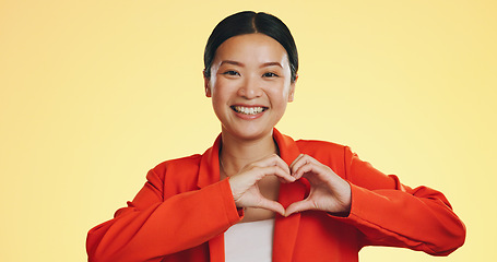 Image showing Smile, heart in hands and face of Asian woman on yellow background for love, support and charity mockup. Emoji, hand gesture and portrait of happy girl in studio with shape for care, trust and kind