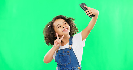 Image showing Phone, selfie and child peace sign in a studio with happiness from profile picture. Isolated, green background and young girl with technology showing a v hand gesture for social media with a smile