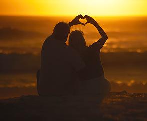 Image showing Silhouette, couple and heart shape at sunset on beach for vacation, holiday or emoji outdoor. Back of man and woman in nature with creative sky, hand icon and ocean for love, art and travel freedom