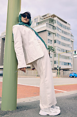 Image showing Portrait, city fashion or religion with a muslim woman leaning against a pole in a cap and scarf for contemporary style. Islam, faith or hijab with a trendy young arab person outside in an urban town