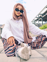 Image showing Portrait, fashion or tradition with a muslim woman outdoor in sunglasses and a scarf for contemporary style. Islam, faith or hijab with a trendy young islamic person sitting outside in modern clothes