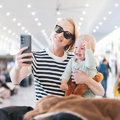 Image showing Mother taking selfie with mobile phone, while traveling with child, holding his infant baby boy at airport, waiting to board a plane. Travel with kids concept.