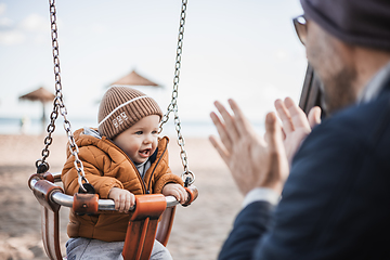 Image showing Father pushing hir cheerful infant baby boy child on a swing on sandy beach playground outdoors on nice sunny cold winter day in Malaga, Spain.