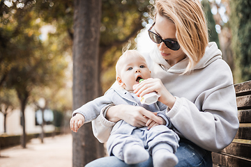 Image showing Mother sitting on bench in urban park, holding her infant baby boy child in her lap and feeding him baby food on nice spring sunny day