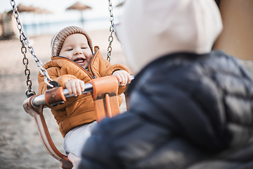 Image showing Mother pushing her cheerful infant baby boy child on a swing on sandy beach playground outdoors on nice sunny cold winter day in Malaga, Spain.