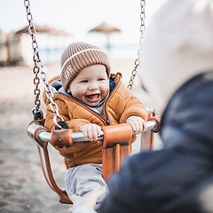 Image showing Mother pushing her cheerful infant baby boy child on a swing on sandy beach playground outdoors on nice sunny cold winter day in Malaga, Spain.