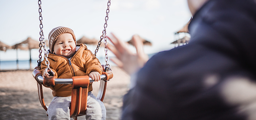 Image showing Mother pushing her cheerful infant baby boy child on a swing on sandy beach playground outdoors on nice sunny cold winter day in Malaga, Spain.