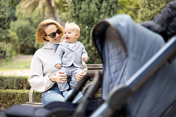 Image showing Mother sitting on bench in urban park, laughing cheerfully, holding her smiling infant baby boy child in her lap having baby stroller parked by their site.