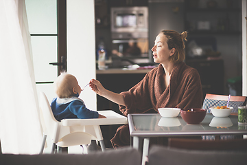 Image showing Cheerful mother wearing bathrope spoon feeding her infant baby boy child sitting in high chair at the dining table in kitchen at home in the morning.