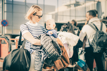 Image showing Motherat travelling with his infant baby boy child. Mom holding travel bag and her infant baby boy child while queuing for bus in front of airport terminal station.