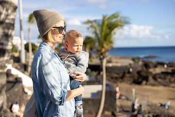 Image showing Fashinable mother holding her infant boy child in lap enjoing panoramic view along palm lined beach promenade of Puerto del Carmen, Lanzarote, Canary islands, Spain.