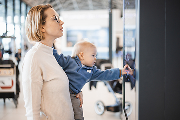 Image showing Mother traveling with child, holding his infant baby boy at airport terminal, checking flight schedule, waiting to board a plane. Travel with kids concept.