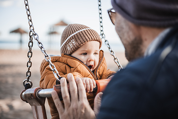 Image showing Father pushing hir cheerful infant baby boy child on a swing on sandy beach playground outdoors on nice sunny cold winter day in Malaga, Spain.