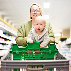 Image showing Mother pushing shopping cart with her infant baby boy child down department aisle in supermarket grocery store. Shopping with kids concept.