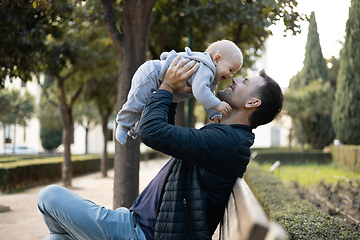 Image showing Father holding and lifting his cute infant baby boy child sitting on wooden bench in urban city park. Dad and son enjoing a pristine moment of happiness, smiling and laughing