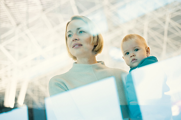 Image showing Thoughtful young mother looking trough window holding his infant baby boy child while waiting to board an airplane at airport terminal departure gates. Travel with baby concept.