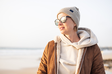 Image showing Portrait of young stylish woman wearing brown padded jacket, hoodie, wool cap and sunglasses on long sandy beach in spring.