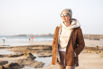 Image showing Portrait of young stylish woman wearing brown padded jacket, hoodie, wool cap and sunglasses on long sandy beach in spring.