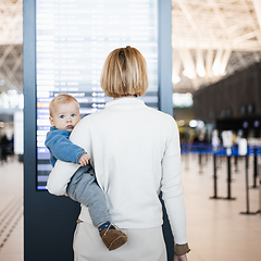 Image showing Mother traveling with child, holding his infant baby boy at airport terminal, checking flight schedule, waiting to board a plane. Travel with kids concept.