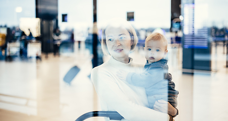 Image showing Thoughtful young mother looking trough window holding his infant baby boy child while waiting to board an airplane at airport terminal departure gates. Travel with baby concept.