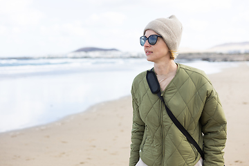 Image showing Portrait of young stylish woman wearing green padded jacket, hoodie, wool cap and sunglasses on long sandy beach in spring.