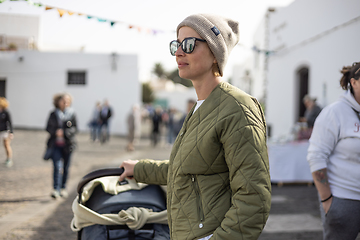 Image showing Portrait of young stylish woman wearing green padded jacket, hoodie, wool cap and sunglasses pershing stroller down cobled street of touristic town of Tequise, Lanyarote, Spain.