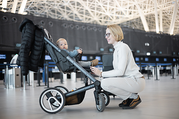 Image showing Motherat interacting with her infant baby boy child in stroller while travelling at airport terminal station. Travel with child concept.