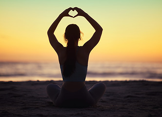 Image showing Woman yoga, heart hand and shadow at sunset at beach and ocean with peace and zen hands sign. Silhouette, nature and sea on a vacation with calm meditation and love emoji gesture outdoor at sunrise