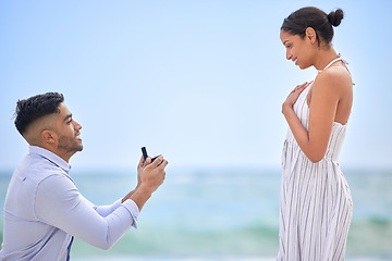 Image showing Engagement, proposal and romance with a couple on the beach for a milestone on a blue sky background. Love, summer or nature with a man down on one knee to ask his girlfriend the marriage question