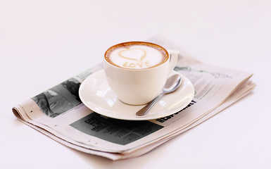 Image showing Coffee cup, cappuccino and newspaper with heart in foam on a table with no people in a restaurant. Cafe drink, love writing and foamy beverage art with milk in a latte and a closeup of mug and spoon