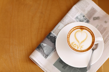 Image showing Coffee, cappuccino and heart in foam with newspaper on a table with no people in a restaurant. Cafe drink, love writing and foamy beverage art with milk in a cafe with a closeup of mug and mockup