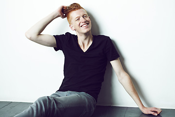 Image showing Portrait, ginger and man sitting on a floor with hand on head or relax or smile for fashion and against white wall. Happy, unique style and excited male model with red hair laugh for fun in studio