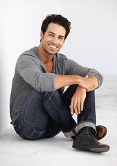 Image showing Floor, portrait and happy man isolated on a wall background with casual clothes, confidence and relax. Handsome model or Portugal person smile and sitting on ground in minimal room with cool fashion