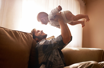 Image showing Dad, funny and lifting baby on sofa in home living room, playing or bonding together. Laughing, care and father holding infant, newborn or child on couch in lounge, having fun and enjoying time.