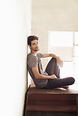 Image showing Portrait, relax and stairs with a man sitting by a window on the landing of a staircase while alone in his home. Happy, content and lifestyle with a handsome young male person laid-back in a house