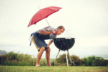 Image showing Umbrella, rain and a man outdoor to barbecue food for cooking or insurance in the winter season. Storm, weather and grill with a male person getting wet while trying to bbq on a grass lawn or garden