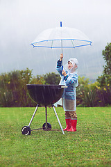 Image showing Rain, umbrella and a boy covering a barbecue outdoor on a field while cooking food alone in winter. Raincoat, kids and insurance with a young male child trying to protect a bbq during stormy weather