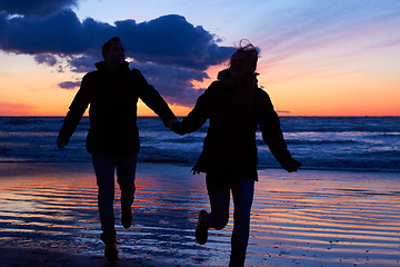 Image showing Sunset, silhouette and couple running at beach on romantic date, vacation or holiday in nature. Man and woman holding hands with love and care on travel, adventure or trip with clouds, sky and ocean